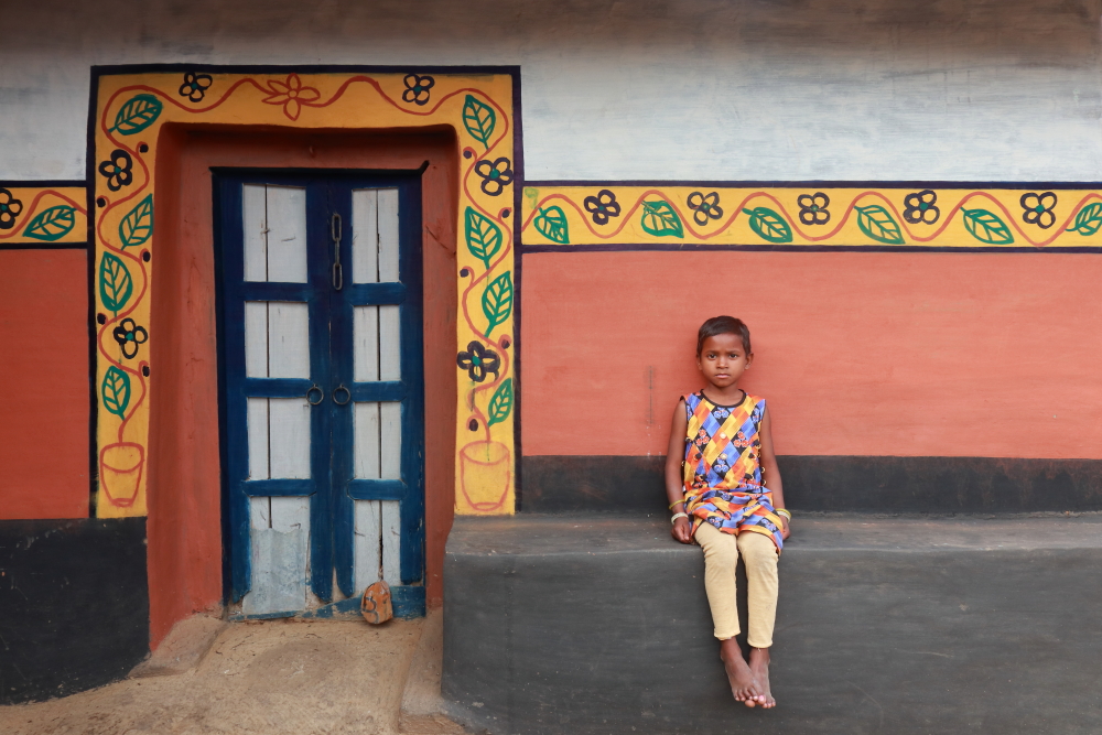 A portrait of a pretty girl from a tribal village in Purulia sitting in front of her beautifully painted home decorated with designs of flowers and leaves.