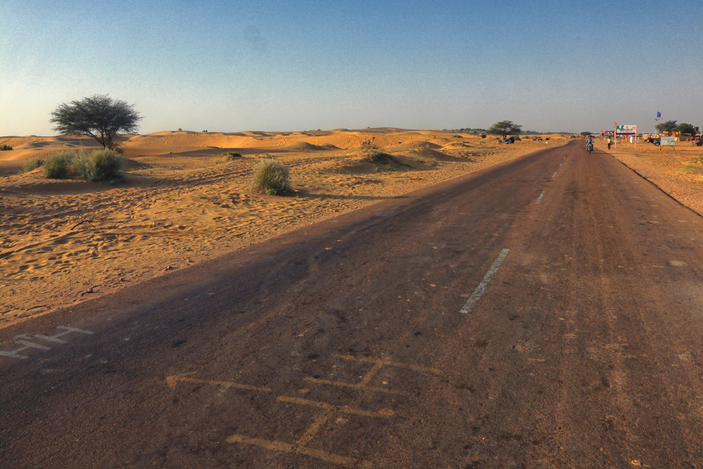 An image of Jaisalmer-Sam highway. The landscape beside the highway is so beautiful, endless stretches of desert in both sides with intermittent dunes and mostly covered with shrubs. There are occasional small villages beside the main road where a narrow road cuts away from the main highway.